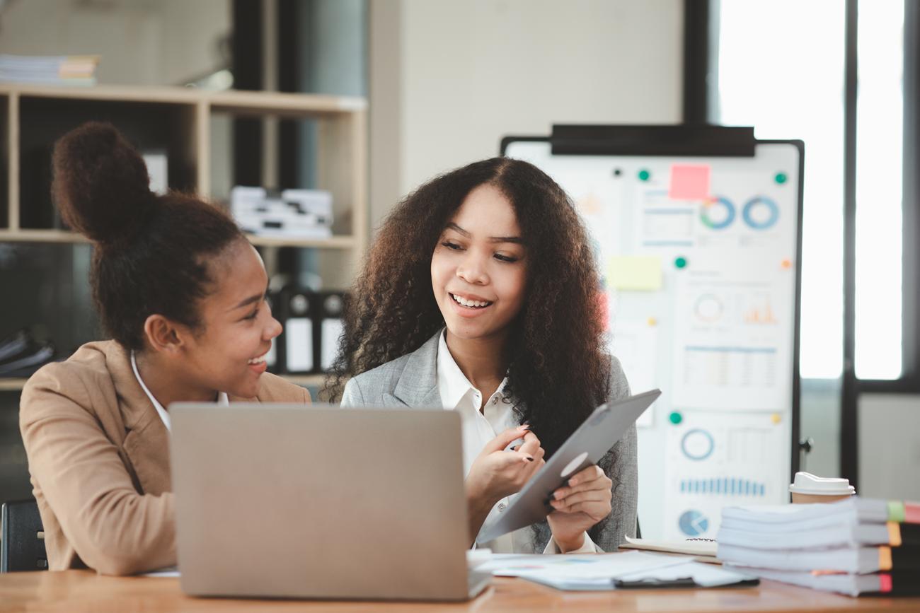 Mulheres jovens em trajes profissionais, sentadas à mesa com um laptop.