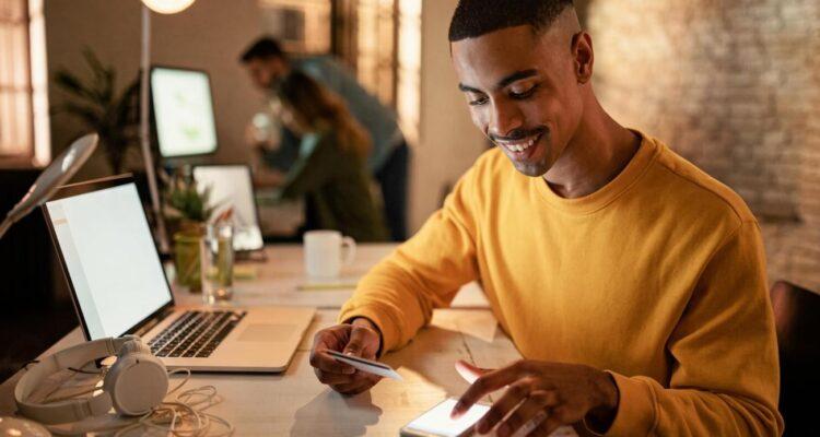 Homem sorridente usando smartphone e laptop.